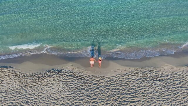 Couple of man and woman stroll hand in hand on the pristine sand of Simos Beach, their footprints tracing patterns as waves gently lap at the shore. The beauty of Elafonisos Greece