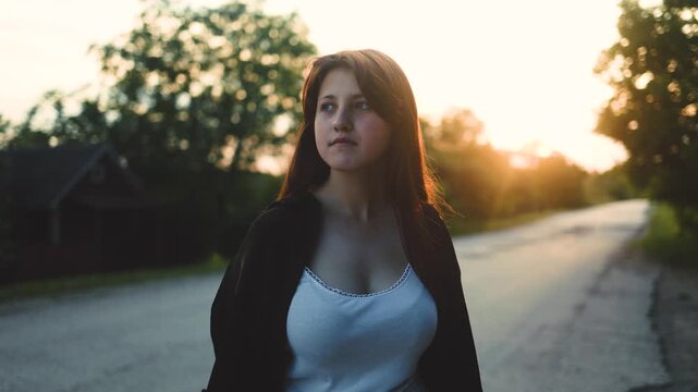 A thoughtful young woman walks along a countryside road during a warm golden sunset, looking ahead. Sunset, nature, travel, calm, journey, serenity, evening, freedom, future, exploration.