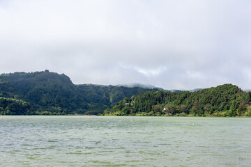 Fototapeta premium Furnas Lake surrounded by lush greenery and hills. Sao Miguel Island in the Azores, Portugal. Volcanic landscape with calm water and cloudy sky.