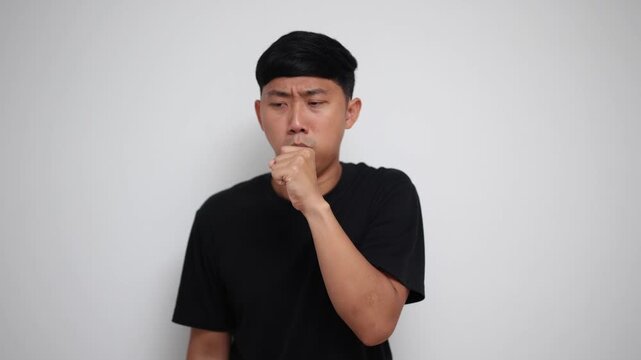young Asian man in a black t-shirt coughing heavily while holding his stomach on a white background