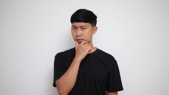 Asian man in a black shirt touches his chin and looks around thoughtfully on white background