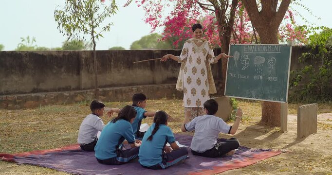 Indian young adult female hold stick pointing words talk speak teach to girl boy rural area place happy teen child group wear uniform sitting mat look board learn do follow tutor basic step at class