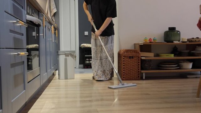 A teenager pushes a flat mop across the kitchen floor near storage shelves during a daily chore.