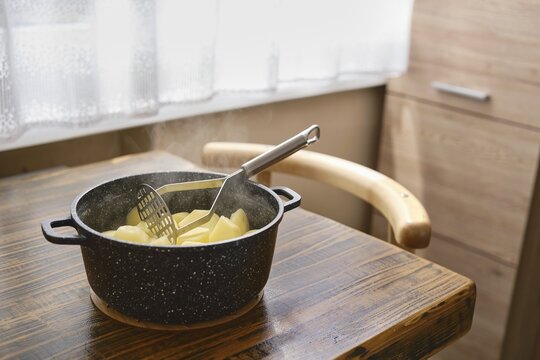 A pot filled with boiled potatoes on a rustic wooden table in a cozy kitchen. Steam rises from the pot, creating a warm and inviting atmosphere. Natural light filters through a nearby window