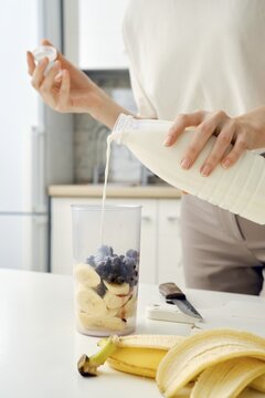 Unrecognizable woman pours milk from bottle into bowl of blender to make a smoothie with black currant