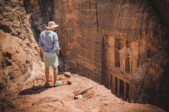 Tourist with sun hat stands on rocks and looks from above into the gorge Siq, Pharaoh's treasure house carved into rock, facade of the treasure house Al-Khazneh, Khazne Faraun, mausoleum in the Nabataean city Petra, near Wadi Musa, Jordan