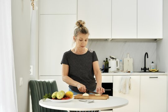 A woman is chopping an onion on a wooden cutting board in a kitchen filled with natural light. Fresh vegetables are arranged on the table, highlighting her cooking activity