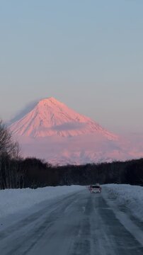 Majestic snow-covered volcano mountain under the first rays of sunrise, with dramatic light illuminating the peaks. A scenic road leads toward the mountain with a car in motion, creating a sense of jo