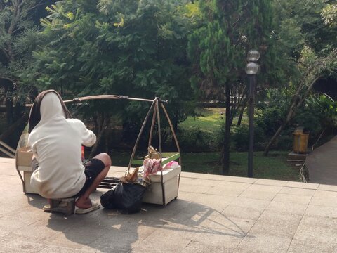 A traditional sate vendor seen from behind sitting in front of his merchandise basket in a city park in Bogor, Indonesia