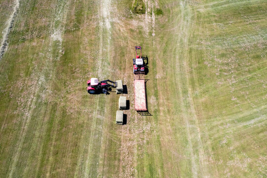Aerial view of two red tractors loading rectangular hay bales onto a trailer in a green agricultural field in Chavannaz, Auvergne-Rhone-Alpes, France.