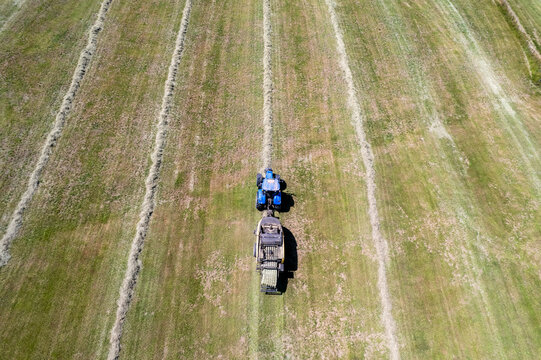 Aerial view of a blue tractor pulling a hay baler through a green field with parallel windrows of cut grass in Chavannaz, Auvergne-Rhone-Alpes, France.