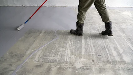 Construction worker applying gray floor coating to concrete surface. Close up of legs and boots during renovation. Industrial flooring and maintenance © Ruslan