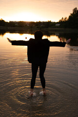 Fototapeta premium silhouetted figure wading barefoot into a shallow lake at golden sunset, arms extended in a peaceful, contemplative pose; warm orange-pink sky reflected in rippling water, dark treeline on the horizon