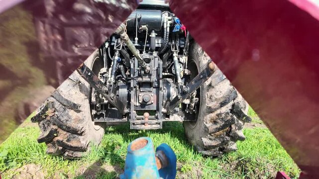 A modern blue agricultural tractor is viewed from beneath a metal frame used for rear attachment. The tractor reverses toward the coupling device, lowering hydraulic arms and aligning the rear hitch