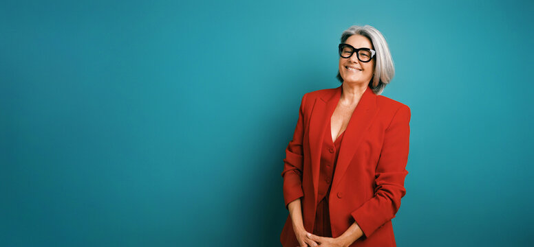 Senior businesswoman in red blazer and stylish glasses expressing confidence and joy, standing on a teal studio backdrop