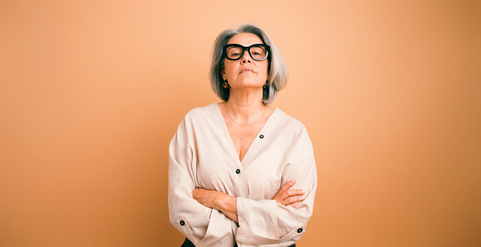 Senior woman wearing glasses and casual clothing, confidently posing with arms crossed on an orange background