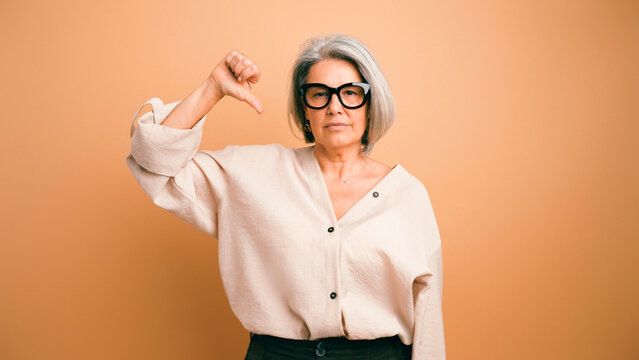 Senior woman with gray hair and glasses showing a thumbs down gesture, expressing dislike, dissatisfaction, and rejection