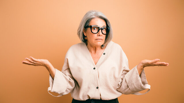 Senior woman shrugging, having a confused and uncertain expression, standing against an orange background