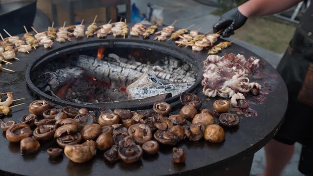 Champignon mushrooms and seafood are grilled on grill. Male hands in gloves turn octopuses over with barbecue tongs. Black flat grill, round shape, with a hole with a fire in center.