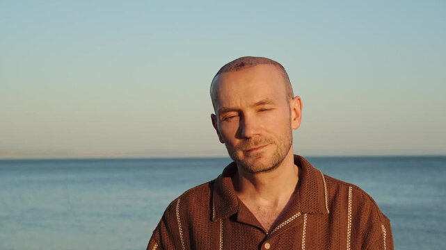 A portrait of a calm and content Caucasian man in a brown polo shirt, showing a healing scalp with subtle scabs after a recent hair transplant procedure against a sunset ocean background