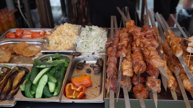 The street cafe's counter offers customers a selection of meat and vegetables grilled over charcoal, along with fresh vegetables. Street food kebab and vegetable counter. Pork skewers.