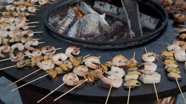 Appetizing seafood, shrimp, and mussels are grilled on skewers on a hot outdoor barbecue. Black flat grill, round shape, with a hole with a fire in center. 