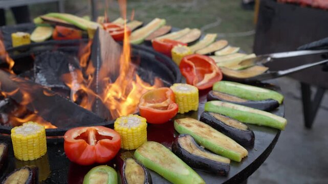 Black flat grill, round shape, with a hole with a fire in center. Vegetables, eggplant, zucchini, corn, and bell peppers are grilled on grill. Male hands in gloves turn food over with barbecue tongs.