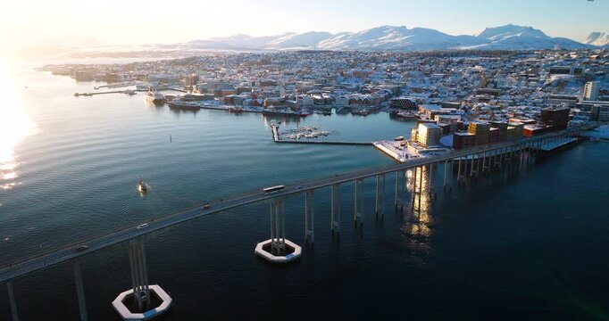 Hermes II boat sailing near Tromso bridge and snow covered city in winter sunlight in Tromso, Norway. Aerial drone panoramic view