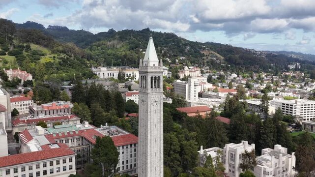Sather Tower rises above UC Berkeley campus with red-tiled buildings and lush green lawns as dense East Bay neighborhoods reach toward San Francisco Bay, California, USA. Aerial drone orbit shot.