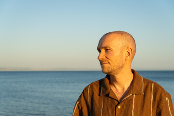 A side profile portrait of a calm and serene Caucasian man looking towards the ocean, showing a healing scalp with subtle scabs after a recent hair transplant procedure at sunset © maxterram