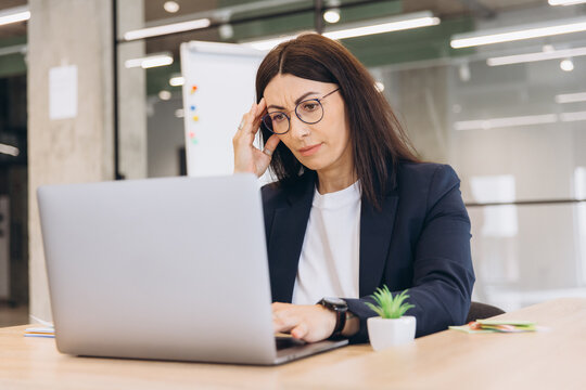 Stressed businesswoman touching her head, suffering from a headache while working on a laptop at her office desk