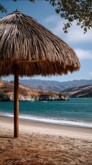 Fototapeta premium Low angle view of a large thatched umbrella on a beach with turquoise ocean and distant mountains under a partly cloudy sky