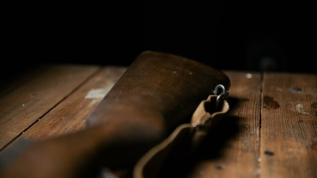 The dark wooden buttstock of a vintage military rifle rests on a rustic wooden table surface next to a tan fabric sling captured in a shallow depth of field shot emphasizing the aged material.