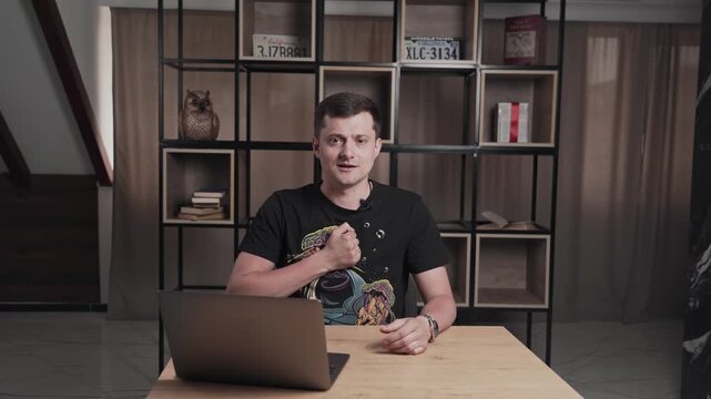 Man in t-shirt in front of laptop talking and punch in the chest. Shelf with books on background. Interview concept.