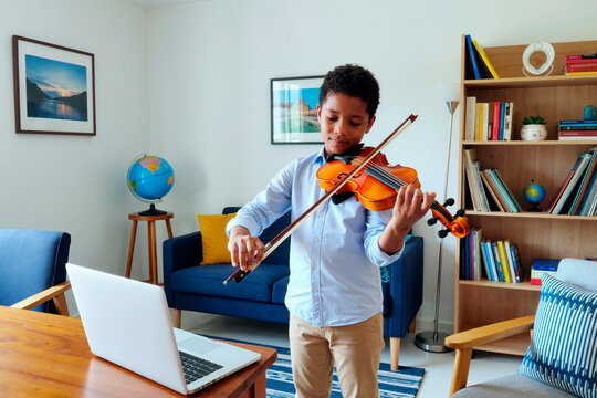 Black boy playing violin beside laptop during online music lesson at home, practicing classical instrument skills. Ideal for remote education, homeschooling, arts training