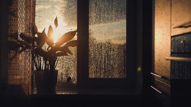 Bright sunset light shines through a rainy window with wet drops on a glass pane in a cozy dark room. Sunset, rain, window, glass, drops, cozy, light, mood, warmth, shadow, evening.