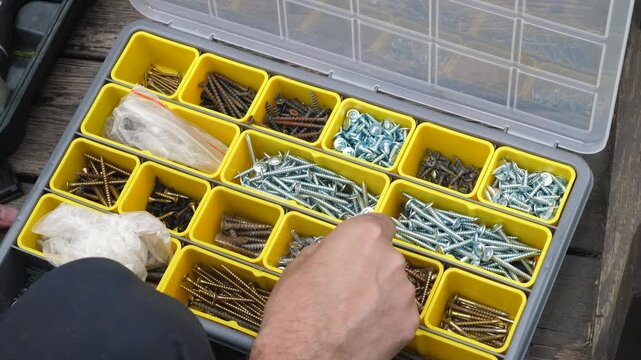 A man looks for the right fasteners in a plastic organizer with removable compartments. The craftsman selects metal screws and bolts from a toolbox during repairs.
