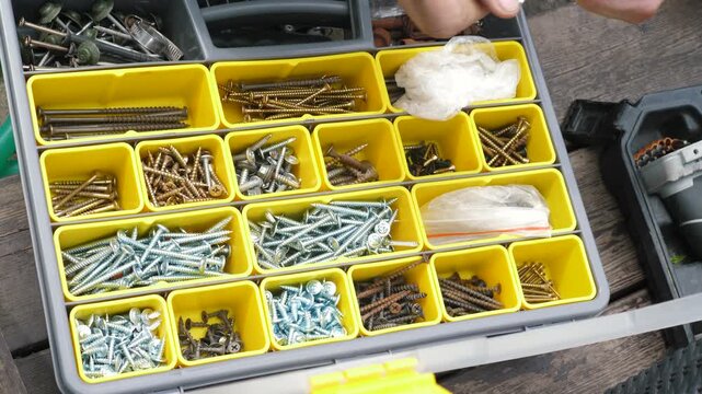 Close-up of male hands choosing the right fasteners from a plastic organizer case. A builder sorts metal screws, bolts and dowels in the yellow compartments of a toolbox