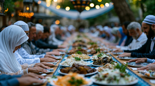 Long communal table set outdoors in a courtyard, dozens of community members seated together at Ramadan iftar, hands extended in prayer just before the evening meal, lanterns and draped fabric overh