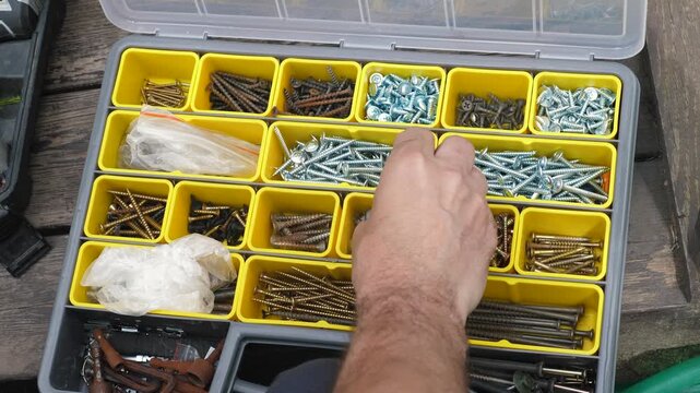 Over the shoulder view of a craftsman choosing the necessary screws from an open toolbox. A man searches through galvanized screws and bolts in the yellow sections of a plastic organizer