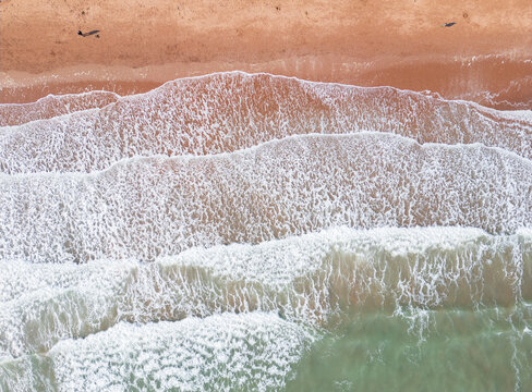Aerial view of Preston Sands beach with white waves crashing on the reddish sand and two people walking along the shore in Paignton, Devon, United Kingdom.