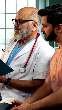 Indian male doctor consulting male patient with medical report as experienced physician advises young man inside modern Indian clinic OPD healthcare consultation