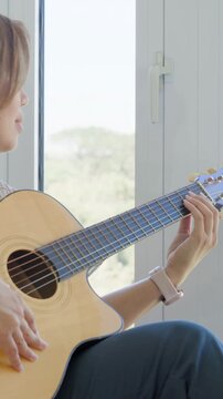 Vertical video: Panning camera revealing woman singing, strumming guitar by window, practicing song