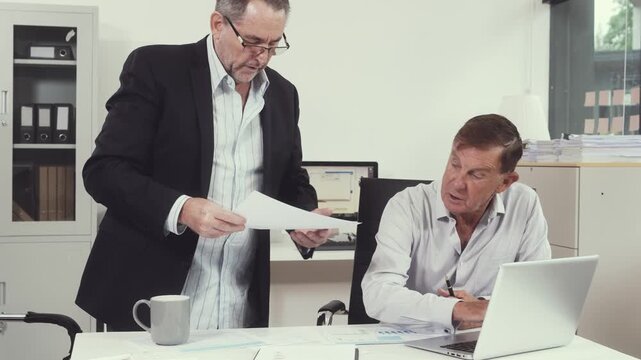 Two businessmen sit at a desk in a bright office, reviewing papers and discussing details during a professional meeting with laptop, tablet and window view.