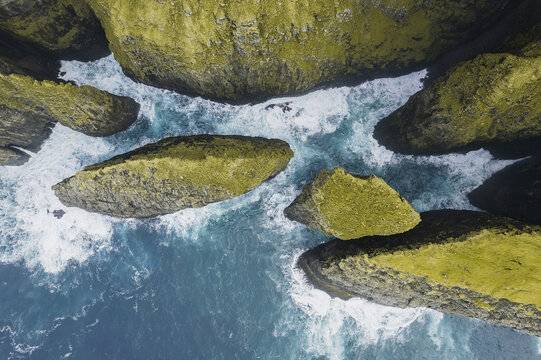 Aerial view of steep mossy green cliffs and sea stacks surrounded by crashing white waves in the blue ocean Faroe Islands.