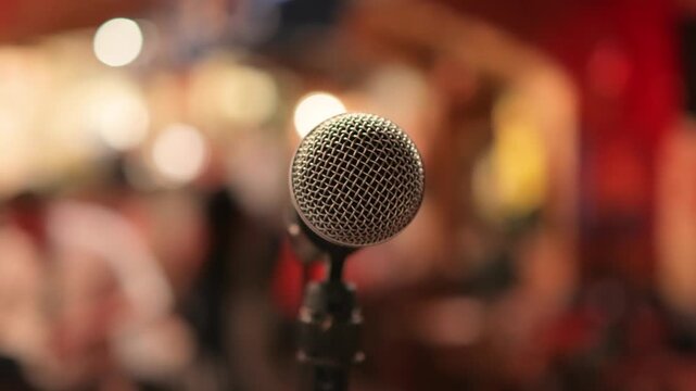 Close up shot of microphone on stand in dimly lit venue, audience blurred in background, live performance setup.