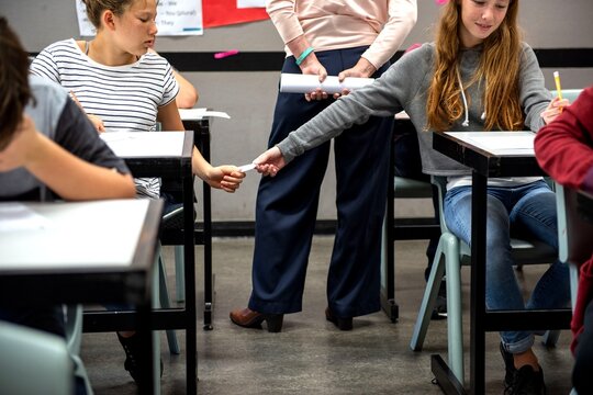 Students passing notes in class while a teacher stands nearby.