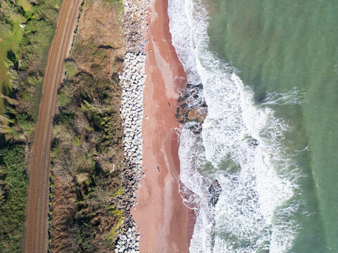 Aerial view of Hollicombe beach featuring red sand, a coastal railway track, and waves crashing against a rocky sea wall in Torquay, Devon, United Kingdom.