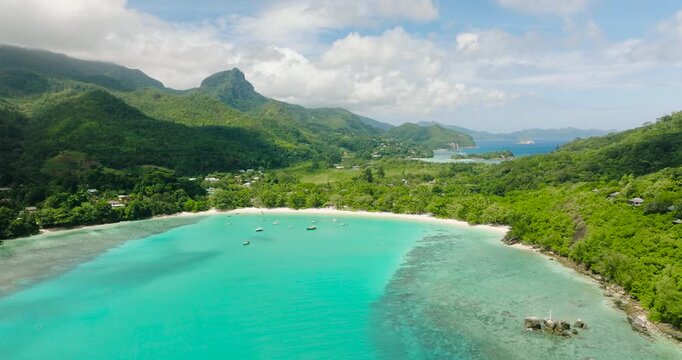 A calm turquoise bay with anchored boats, surrounded by lush green hills and tropical vegetation. Port Launay. Seychelles, Mahe.