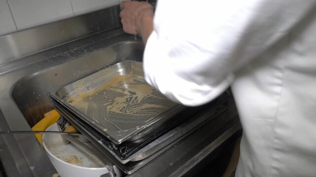 A kitchen worker scrubs large baking trays in the sink after a busy catering event, an essential cleaning task that keeps the restaurant kitchen running smoothly between services.